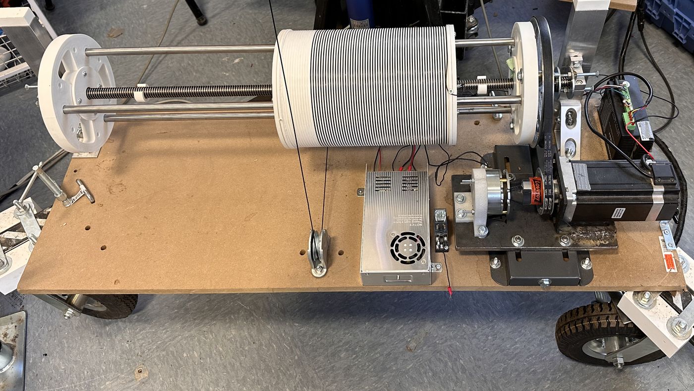 An early test iteration of a spool unit on its cart: white 3D-printed spool and end supports on a lead screw with three steel rods, the large pulley and belt, an old stepper motor with its brake, and the power supply